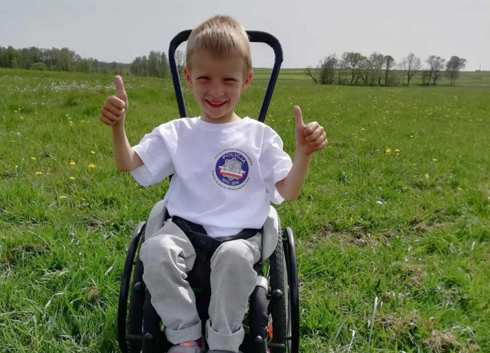 Zdjęcie przedstawia 6-letniego Jasia na wózku inwalidzkim trzymającego kciuki w górę, w  tle łąka 