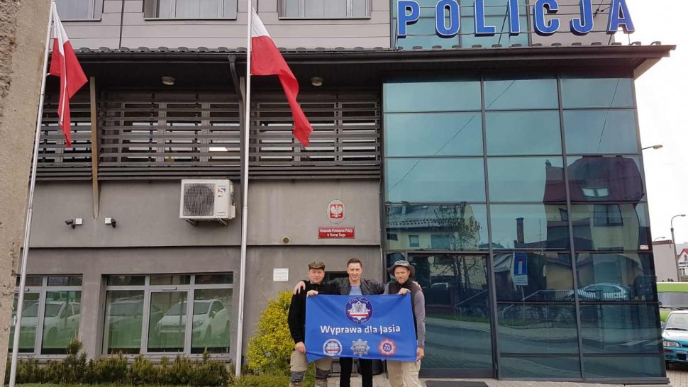 Obrazek przedstawia trzech mężczyzn trzymających flagę z Wyprawy dla Jasia, stojących na tle Komendy Powiatowej Policji w Nowym Targu.