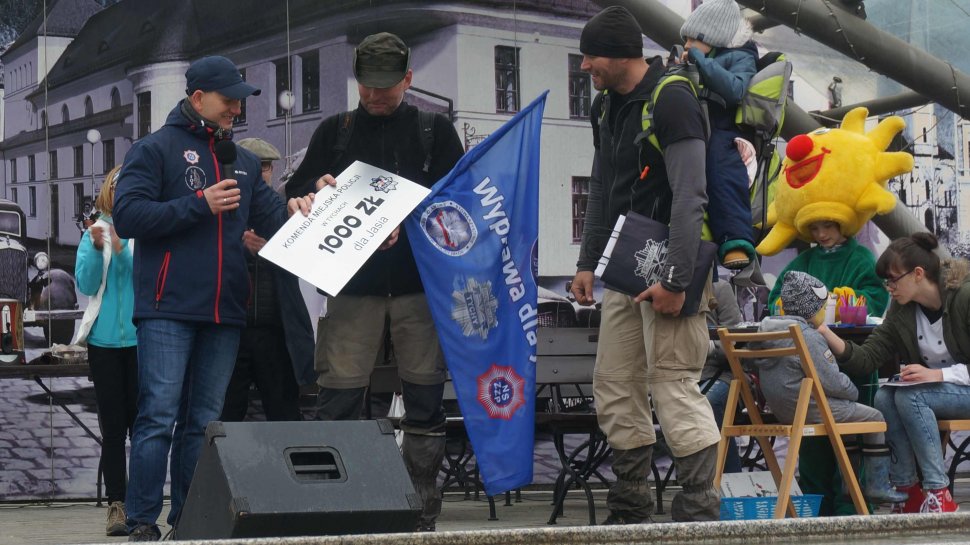 Obrazek przedstawia trzech mężczyzn na scenie, jeden z nich na plecach trzyma w foteliku chłopca, obok niego inny tzryma flagę "Wyprawa dla Jasia i przyjmuje potwierdzenie zapłaty w postaci bonu od stojącego z lewej strony mężczyzny 