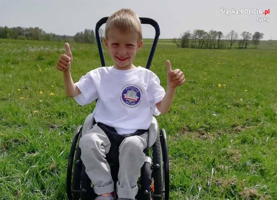 Brazek przedstawia 6-letniego chłopca siedzącego na wózku inwalidzkim i trzymającego kciuki do góry
