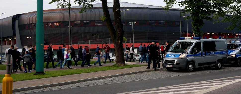 Na obrazku widać stojących przy radiowozie policjantów przed stadionem, na który zmierzają kibice