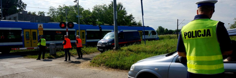 Obrazek przedstawia policjanta stojącego tyłem przy samochodzie, a w tle przejeżdżający pociąg oraz funkcjonariuszy polskich lini kolejowych oraz sokistów