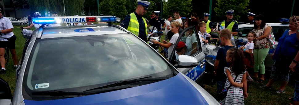 Na zdjęciu widoczny radiowóz, który ma włączone sygnały świetlne obok stoi policjant, a wokół niego uczestnicy festynu