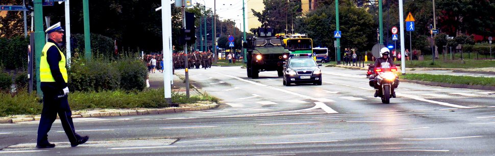 Policjant z drogówki w żółtej kamizelce zabezpiecza skrzyżowanie przez które przechodzi defilada z okazji obchodów Święta Wojska Polskiego, na przodzie jedzie policjant na motocyklu.