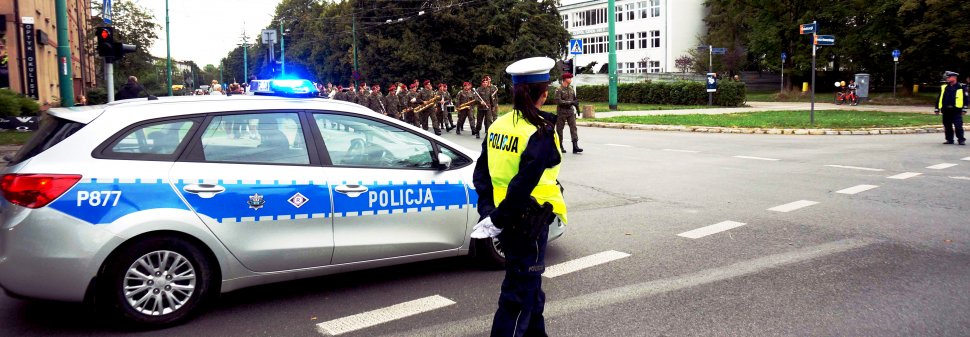 Policjantka wydziału ruchu drogowego, stoi przy radiowozie i zabezpiecza defiladę z okazji obchodów Święta Wojska Polskiego.