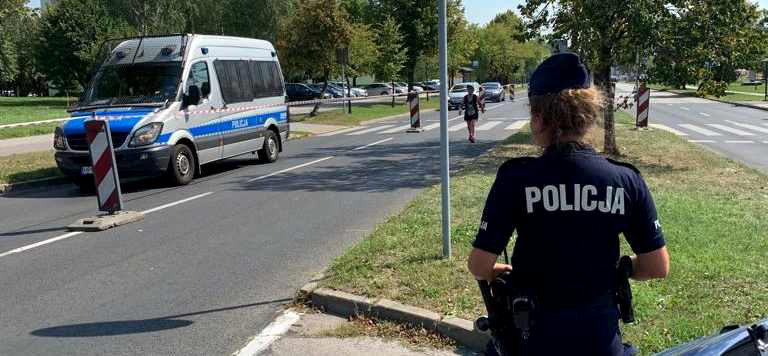 Widoczna policjanta, która stoi tyłem, na przeciwko niej na jezdni zaparkowany radiowóz, widoczny także uczestnik półmaratonu. 