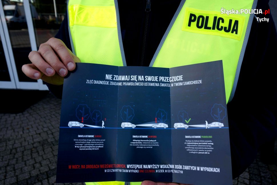 Na zdjęciu policjant w żółtej kamizelce pokazuje do zdjęcia ulotkę dotyczącą kampanii "Twoje światła-nasze bezpieczeństwo". 