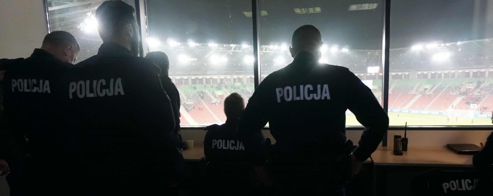 Na zdjęciu pięciu umundurowanych policjantów , którzy stoją przodem do stadionu, gdzie jest widok na murawę, gdzie trwa mecz. 
