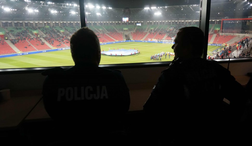 Na zdjęciu dwóch umundurowanych policjantów, siedzą tyłem do zdjęcia, przodem do murawy boiska, gdzie rozpoczyna się mecz. 
