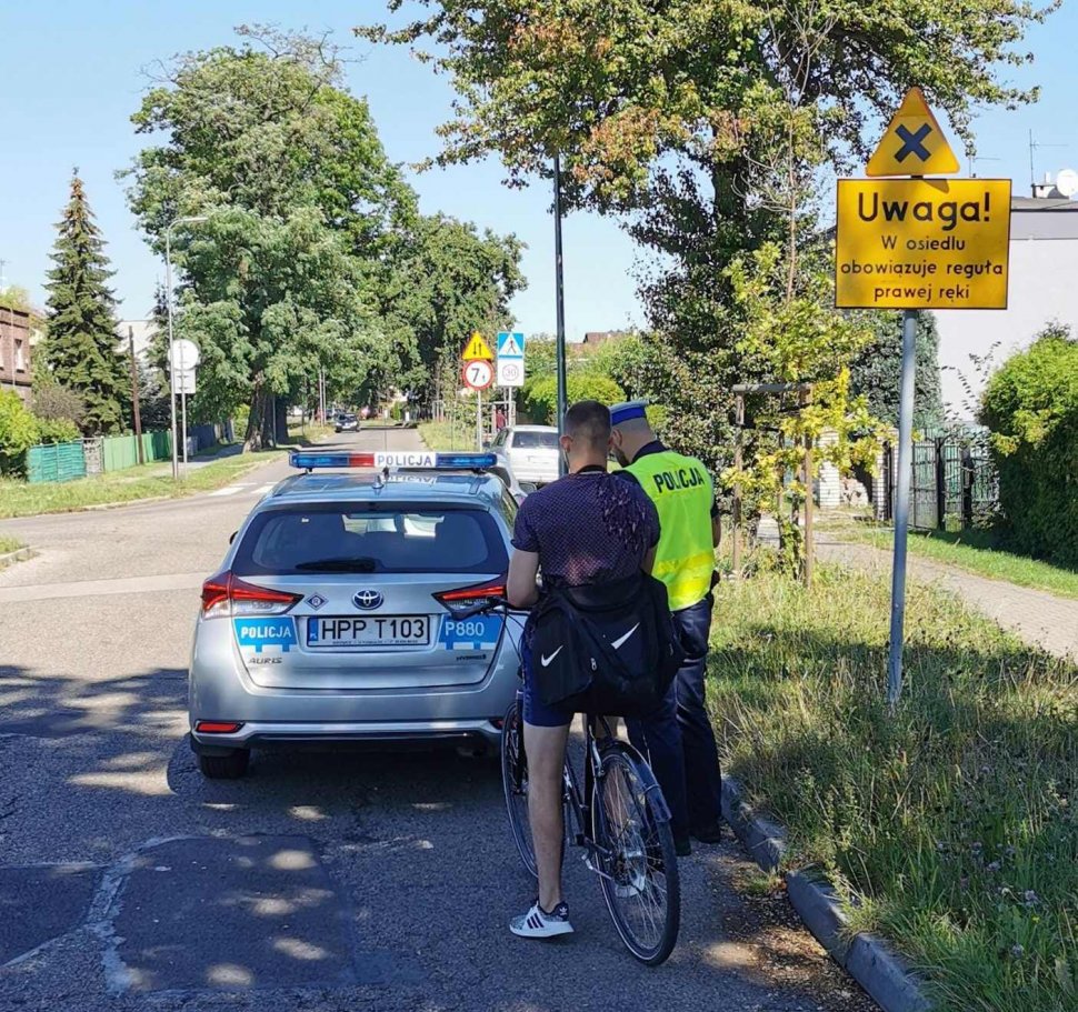 Policjant kontroluje rowerzystę podczas działań pod nazwą "Bezpieczny rower". Przed nimi stoi radiowóz. 