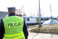 Obrazek przedstawia stojącego &quot;tyłem&quot; policjanta  z przodu widać zamknięty przejazd kolejowy i pędzący pociąg, z prawej strony w tle radiowóz