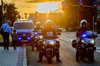 Obrazek przedstawia z przodu trzech policjantów na motocyklach, dalej po lewej stronie radiowóz a obok niego przechodzi mężczyzna z dzieckiem