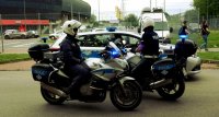 Obrazek przedstawia policjanta na motocyklu, radiowóz, w tle inne osoby.