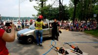 Na zdjęciu widoczny pojazd, oraz ubrana w strój strażaka kobieta, a także strażak, w tle tłumy zgromadzonych dzieci z opiekunami