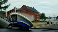 Widok zdjęcia zrobionego wewnątrz z radiowozu, widać na podszybiu czapkę policjanta wydziału ruchu drogowego, w tle idących ministrantów z krzyżem