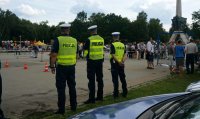 Na zdjęciu trzech policjantów stoi tyłem w kamizelkach odblaskowych podczas zlotu samochodowego, w tle widoczne inne osoby