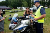 Na zdjęciu widoczny policjant wydziału ruchu drogowego, stojący przy policyjnym motocyklu, na którym siedzi dziewczynka, a ją trzyma mama. Policjant wręczył dziecku opaskę odblaskową oraz kolorowankę.