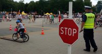 Na zdjęciu widoczny znak drogowy &amp;quot;STOP&amp;quot;, dalej widoczny policjant, który patrzy na dziecko jadące na rowerze po torze przeszkód. Widoczne także inne osoby, które są uczestnikami festynu.