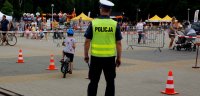Na zdjęciu widoczny policjant, który patrzy na dziecko jadące na rowerze po torze przeszkód. Widoczne także inne osoby, które są uczestnikami festynu.