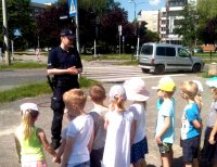 Umundurowany policjant tłumaczy dzieciom jak bezpiecznie przejść przez jezdnię, wszyscy stoją obok przejścia dla pieszych