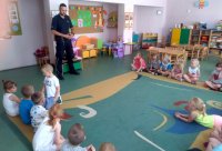 W sali przedszkolnej dzieci siedzą na dywanie, przed nimi stoi umundurowany policjant