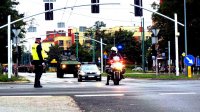 Policjant z drogówki w żółtej kamizelce zabezpiecza skrzyżowanie przez które przechodzi defilada z okazji obchodów Święta Wojska Polskiego, na przodzie jedzie policjant na motocyklu
