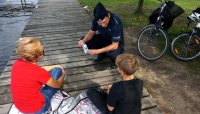 Policjant kuca i rozmawia z chłopcem i jego mamą, którzy wypoczywają nad jeziorem.