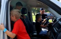 Zdjęcie przedstawia wnętrze samochodu, w którym znajdują się dzieci, za oknem dogląda ich umundurowany policjant.
