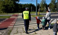 Policjant drogówki w kamizelce odblaskowej przeprowadza przez przejście dla pieszych dzieci, które idą ze swoja opiekunką, która trzyma rękę do góry.