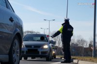 Na zdjęciu widoczny policjant wydziału ruchu drogowego, który zatrzymuje pojazd do kontroli. Za nim widoczne jadące inne samochody.