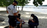 Policjantka siedzi na ławce i rozmawia o bezpieczeństwie nad wodą z kobietą i jej  dzieckiem podczas spotkania profilaktycznego. Obok stoi umundurowany policjant.