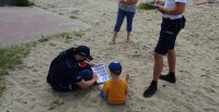 Policjanta siedzi na plaży z chłopcem i pokazuje mu plansze ze znakami ostrzegawczymi związanymi z bezpieczeństwem nad wodą.