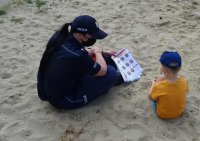 Policjanta siedzi na plaży z chłopcem i pokazuje mu plansze ze znakami ostrzegawczymi związanymi z bezpieczeństwem nad wodą.