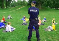 Policyjny profilaktyk rozmawia z przedszkolakami, które siedzą na matach na trawie.