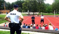 Policjanci stoją na boisku przed grupą przedszkolaków i rozmawiają z nimi o bezpieczeństwie w ramach prowadzonych działań profilaktycznych.