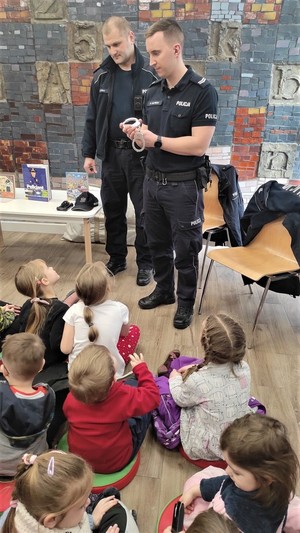 Na zdjęciu dwóch umundurowanych policjantów. Jeden z nich  pokazuje dzieciom kajdanki. Przed policjantami grupa dzieci w wieku przedszkolnym.