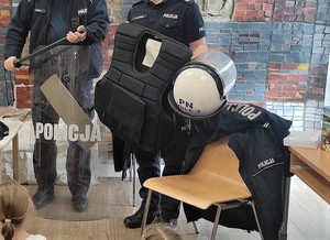 Na zdjęciu mundur policyjny na krześle oraz sprzęt do zabezpieczania imprez: kask, kamizelka, tarcza i pałka.