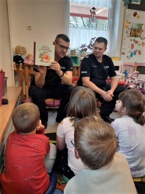 Na zdjęciu dwóch umundurowanych policjantów siedzących przed grupą dzieci. Dzieci siedzą przed policjantami na kolorowym dywanie. Wokół nich zabawki. Jeden z policjantów ma założone okulary i pokazuje dzieciom książkę.