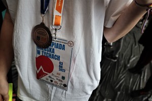 Na zdjęciu osoba w białej koszulce, która ma zawieszony na klatce piersiowej medal oraz legitymację uczestnika olimpiady