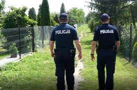Na zdjęciu dwóch umundurowanych policjantów idących drogą. po ich prawej i lewej stronie widoczne są ogródki działkowe.