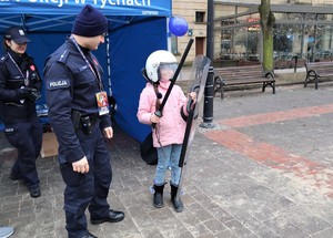 Na zdjęciu policjant oraz dziecko z założonym kaskiem oraz policyjną tarczą i pałką podczas obchodów 32. Finału Wielkiej Orkiestry Świątecznej Pomocy.