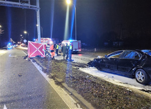 Zdjęcie przedstawia miejsce wypadku drogowego. Widoczny jest rozbity pojazd oraz osoby biorące udział w czynnościach.